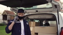 Handsome delivery man checking delivery list while talking on the phone. Delivery man in cap and gloves with phone and documentation in hands outdoor. Trunk with parsels.