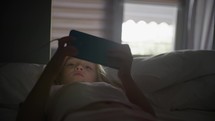 A six-year-old blonde girl, lying on a bed, becomes engrossed with a mobile phone game, colorful light reflections playfully dance across her face, while window light softly fills the background.