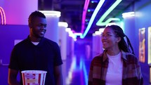 Happy young african american woman and man walking by neon hall with bucket of popcorn, chatting at cinema.