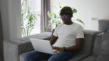 Attractive african American man in glasses sitting on the sofa using laptop at home.