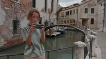 A teenager holds a smartphone near a Venetian canal. A bridge is seen in the background