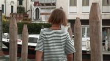 Teenager in striped shirt filmed from behind takes a photo of a scenic Venetian canal with wooden mooring posts