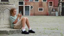 A teenager sits at a Venetian square, deeply engrossed in his smartphone. Dressed in a striped shirt and casual shorts, he seems oblivious to the historic beauty around him