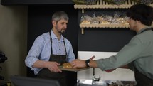 Handsome father in apron passes a tray of pastries to his young son and enters data into the bakery database. Indoor, close up, slow motion.