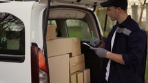 Courier making notes in documents and takes cardboard box package out of delivery car. Courier in uniform, cap and gloves on the way to deliver postal parcel to a client.