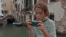 A teenager stands near a Venetian canal, using a smartphone, with historical buildings and a bridge in the background