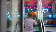 A 12-year-old boy with long hair, seen from behind, throws a basketball into a moving hoop in an arcade game area.