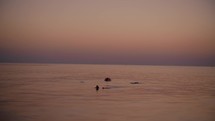 Teen boy submerged to neck in sea, eyes lifted towards the sunset sky, with merely his head, hands, and feet breaking the calm surface, exuding tranquility