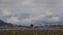 Airplane takes off on runway amidst lush greenery, mountainous backdrop under cloudy sky
