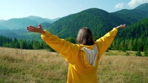 Woman in yellow stands, raises her hands up in front of high Carpathian mountains