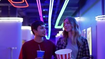 Happy young woman and man walking with bucket of popcorn and drink, chatting at cinema.