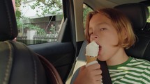 Teenage boy, 12, in a striped tank top, sits in the back passenger seat of a car, eating ice cream. 