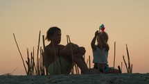 Siblings on the beach construct a bamboo enclosure, with the girl adding a colorful wind spinner to a bamboo straw, enveloped by the gentle sunset glow