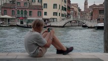 Focus starts on a gondolier rowing in the Venice canal, then pulls to a teenager in a striped shirt sitting and playing on his mobile phone