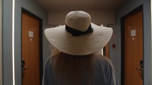 Steadicam shot of a blonde woman walking away in a hotel hallway, her wide-brimmed hat in focus