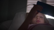 A six-year-old blonde girl lies on the bed, engaging with a mobile phone, while light from a window illuminates the background.