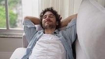 Caucasian man lies down on white leather couch to relax after work. Young white guy feels good smiling at home in slow motion
