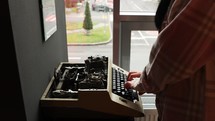 A person typing on a vintage manual typewriter by the window with vehicles passing by outside. 