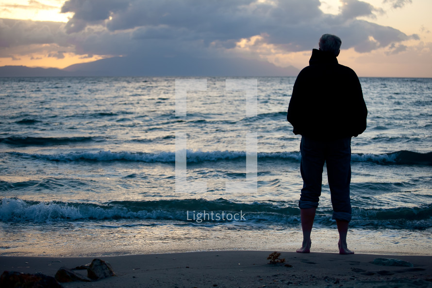 Silhouette of man in jacket with jeans rolled up standing on beach with waves rolling in and cloudy sky at sunset.