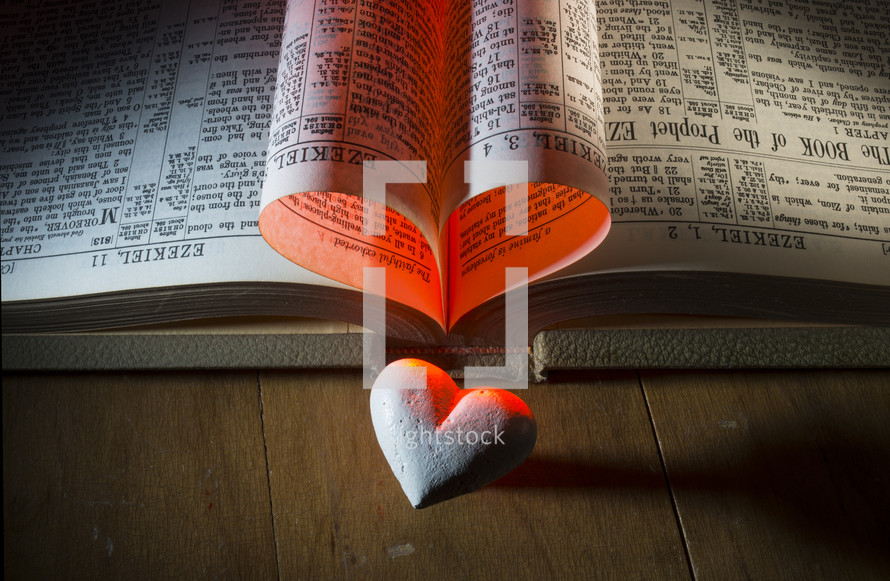 Pages of Bible folded into a heart with a stone heart on a wooden table.