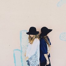 Two women in hats stand back to back against a white wall.