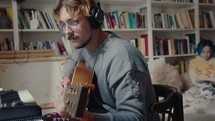 Young musician in headphones drinking red wine and playing the guitar at home recording studio, girlfriend reading a book on bed in the background
