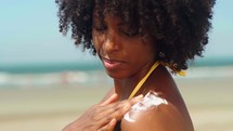 Close up of a happy smiling young black woman is applying a sunscreen or sun tanning lotion on a shoulder to take care of her skin on a seaside beach during holidays vacation.