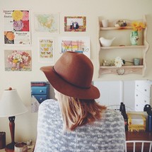 A woman in a hat sits in a room with pictures on the wall.