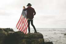 a man with an American flag standing on a rock at a shore 