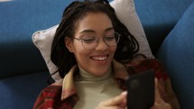 Happy African American teen relaxing on sofa at home with smartphone.
