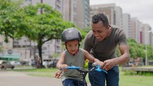 Happy latin father helping smiling boy to ride bike in city park.
