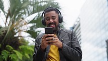 Cheerful young man walking on city street with headphones and modern smartphone in hands. Black man reading some good and smiling. Technology for lifestyles.
