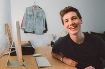 smiling face of a young man sitting at a computer desk 