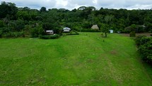 Aerial shot drone flying right while camera pans left over open green field of indigenous village as hikers exit the jungle and enter the village in middle of Amazon rainforest