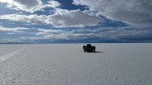 Aerial shot drone flies low to ground and orbits to right around group of tourists in jeep in middle of bright white salt flats contrasting with blue sky