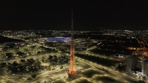 Drone orbits around orange TV Tower as it changes to red, with blue Arena BRB Mane Garrincha in background and lit up Ferris Wheel in Castelinho do Parque da Cidade in Brasilia, Brazil