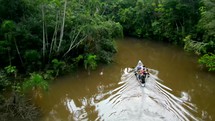 Aerial shot drone hovering as canoe full of people paddling down brown river in middle of Amazon rainforest appear in frame and exit behind trees