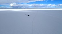 Aerial shot drone lowers over expansive salt flats with only one jeep in the middle of nowhere contrasted by bright blue sky with white and gray clouds