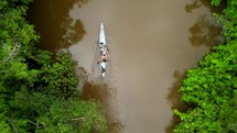 Aerial shot drone flying up as camera points straight down following canoe full of people paddling down brown river in middle of Amazon rainforest
