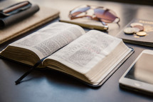 wallet, journal, open Bible, iPhone, coins, sunglasses, pen, and passport on a table 