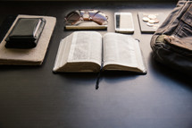 wallet, journal, open Bible, pen, coins, bag, iPhone, sunglasses, and passport on a table 