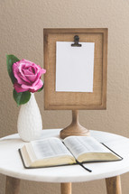 clipboard on a stand, blank paper, flower in a vase, and open Bible on a stand 