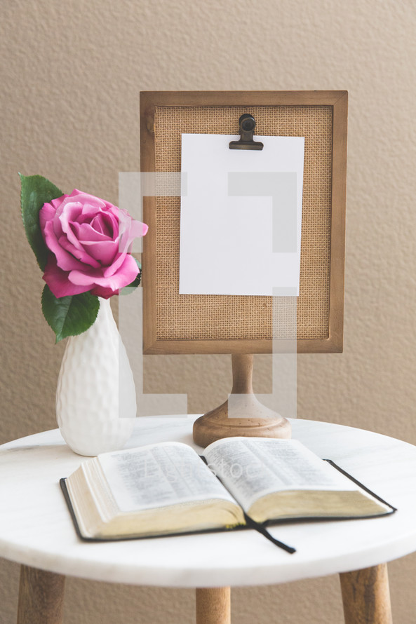 clipboard on a stand, blank paper, flower in a vase, and open Bible on a stand 