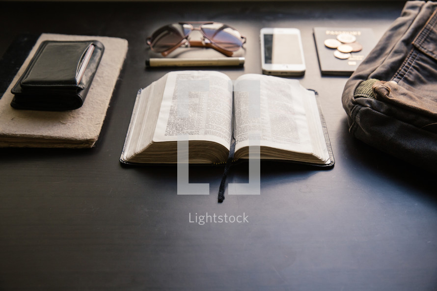 wallet, journal, open Bible, pen, coins, bag, iPhone, sunglasses, and passport on a table 