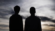 Silhouette of two happy, smiling young men, best friends, or Christian brothers standing in nature watching the summer sunset in cinematic slow motion. 