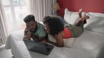 Happy black millennial man and woman using laptop computer at home watching online show lying in bed together. Young adult couple looking at notebook screen in bedroom at home.