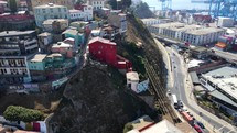 Aerial shot drone slowly orbits to right around red house next to historical funicular next to busy highway and large cranes on loading docks of coastal city on sunny day