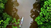 Aerial shot drone lowering with camera pointing down as canoe full of people paddling down brown river in middle of Amazon rainforest flows beneath