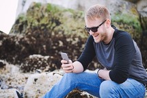 a man sitting on a rock texting on a cellphone 