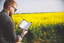 a man standing in a field of yellow flowers reading a Bible 